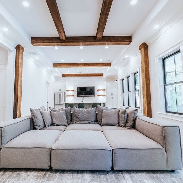 A basement with large dark oak beams and huge gray couch centered in the middle with lots of bright light from the black casement windows with grilles. 