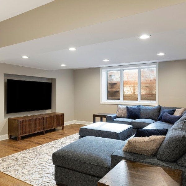 A tan painted basement with a dark oak wood TV stand, a large patterned rug, gray sectional and sliding white windows.