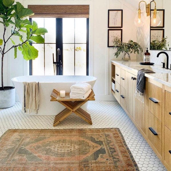 A modern, boho-esque bathroom with white hexagon tiled flooring, light brown vanity, black accents, and white farmhouse tub with a wall of black grille windows behind.  