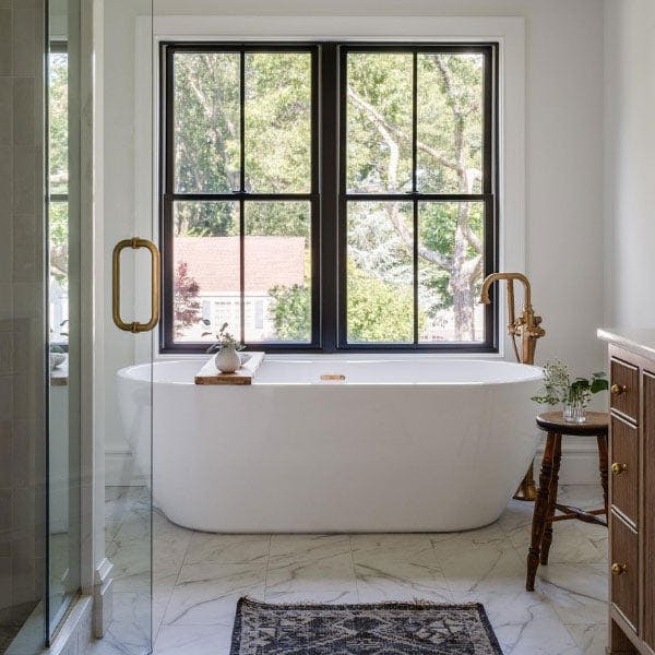 A bright white and modern bathroom with marble granite flooring, gold accent details and large white farmhouse tub is flooded with light from the black windows with grilles installed directly above.