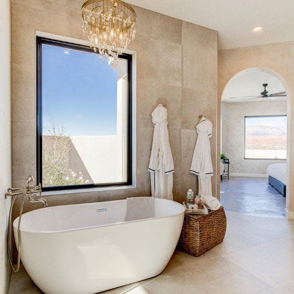 A modern bright tan tiled bathroom with tan tiled flooring, golden hanging chandelier and a black picture window above a large white farmhouse tub.