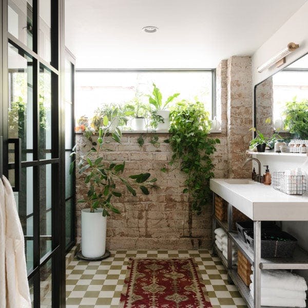 A modern, boho bathroom with white and green checkered tiled flooring, patterned bath matt below a white vanity and rectangle picture window letting in a plethora of light. 