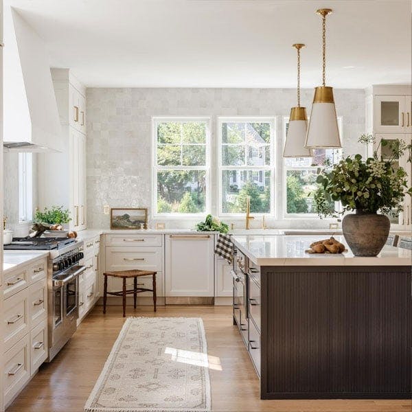 A bright cottage core kitchen with a large center island, white marble countertops, stainless steel appliances and white cabinetry is flooded with light from the white double hung windows with grilles.