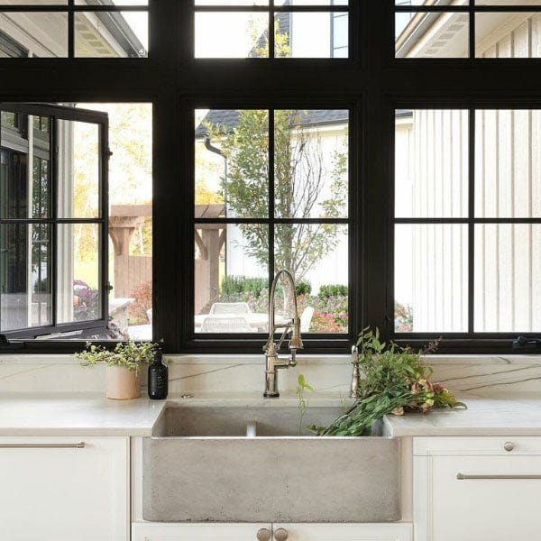 A kitchen with marble countertops, a steel farmhouse sink and white cabinetry is bright with light from the black windows with grilles perched above the sink.