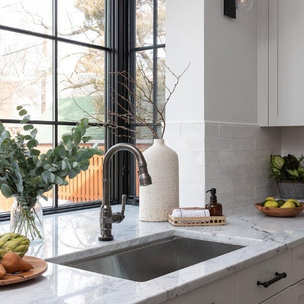 A bright white and modern kitchen with marble granite countertops, stainless steel appliances and white cabinetry is flooded with light from the black windows with grilles perched above the sink.
