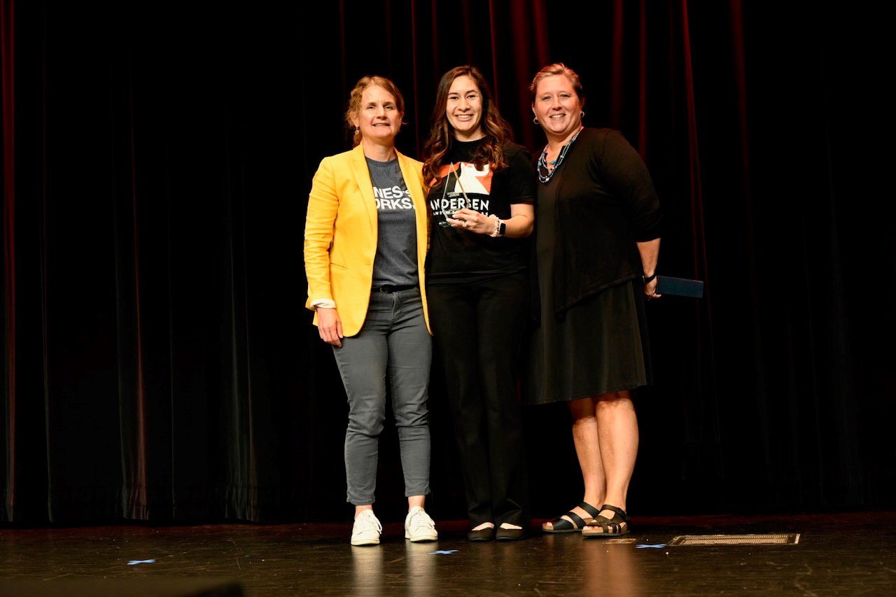 3 women standing on a stage one wearing an Andersen shirt and one wearing a Genesys shirt