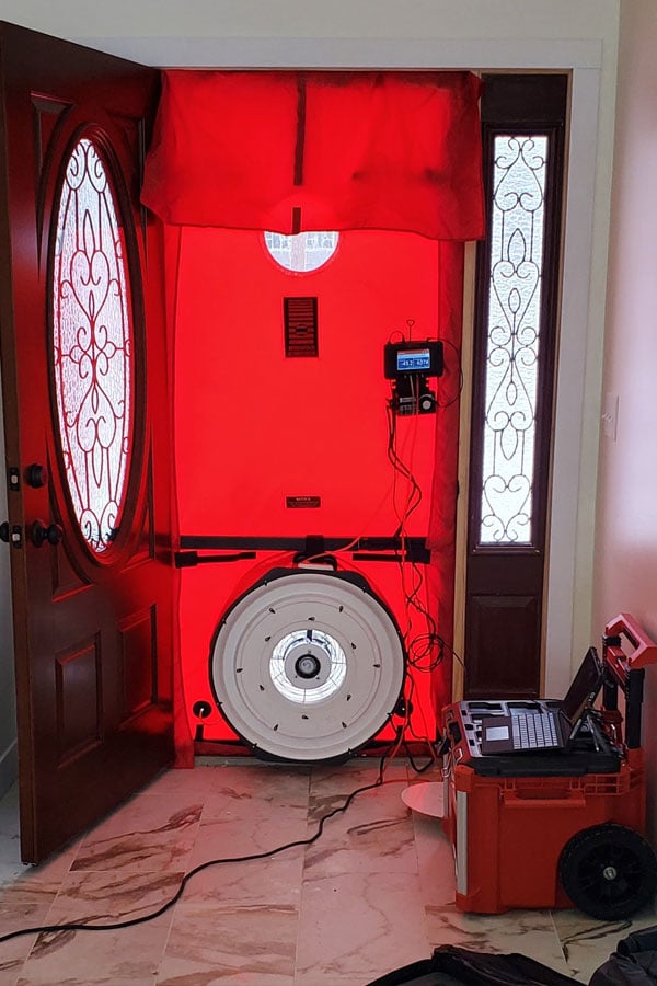 A blower door setup installed in a home's front doorway, with a red canvas and a large circular fan at the bottom. An attached control panel and laptop are visible on the right side, used to measure the building's air leakage. The ornate glass panels on either side of the doorway indicate it is inside a house.
