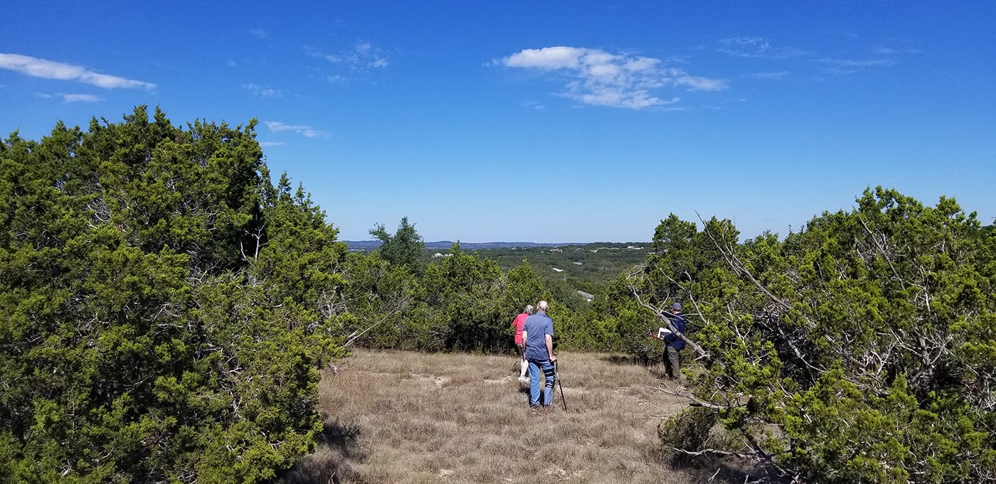 Three people scout a site with brown grass and bushy trees overlooking a wide-open view of the horizon.