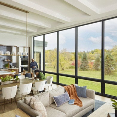 A couple enjoy their kitchen, which features a monumental wall of windows overlooking trees and a pond.