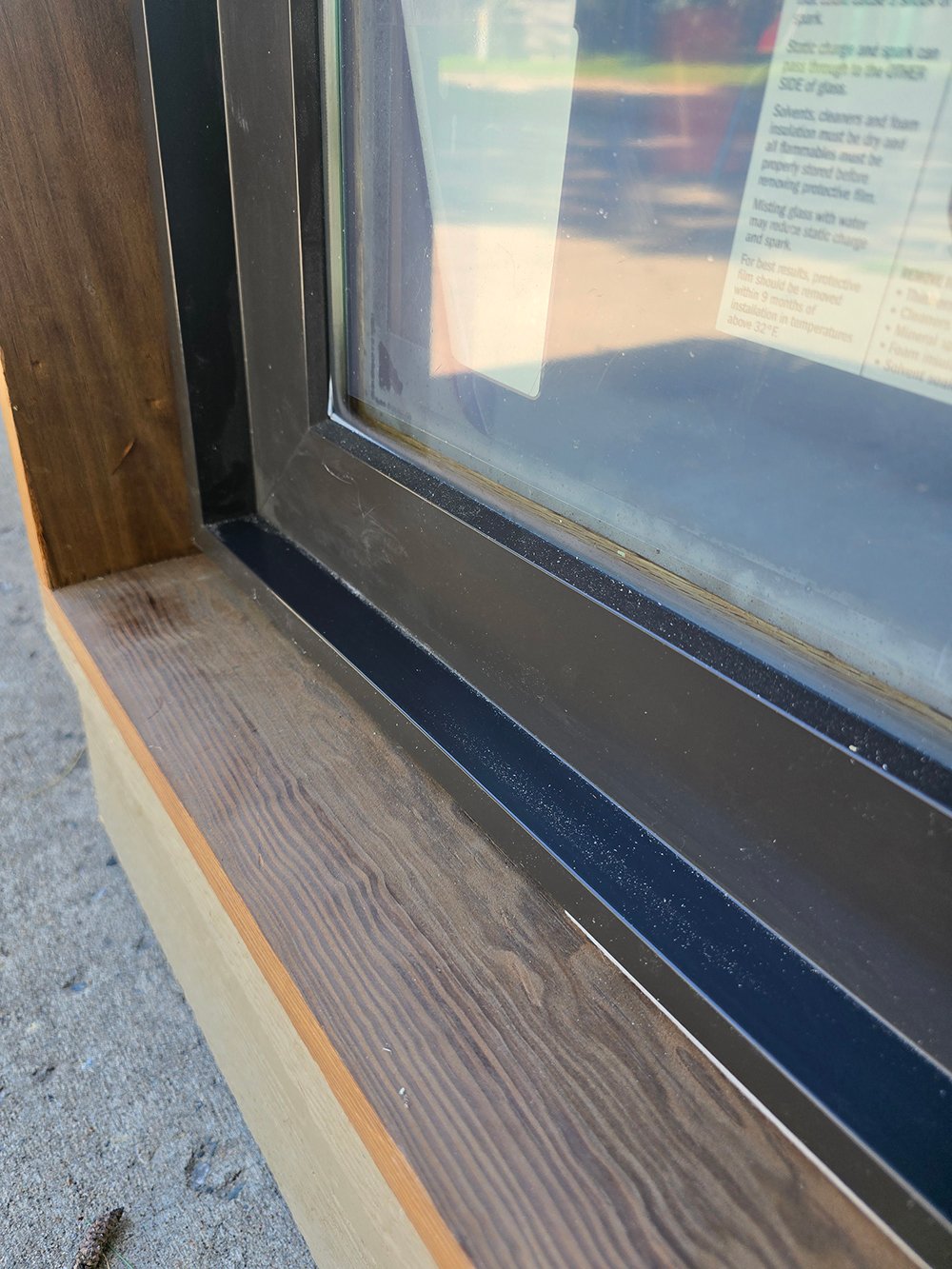 A black middie window with Andersen stickers sits about two-thirds of the way inside its wood rough opening.