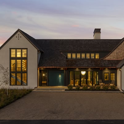 A modern Tudor style home with large black windows lit up from inside.