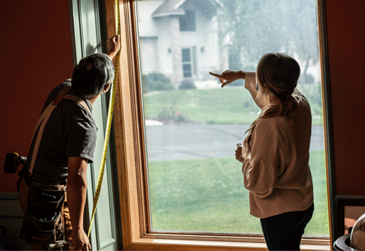 A contractor measures a window while talking with a homeowner.