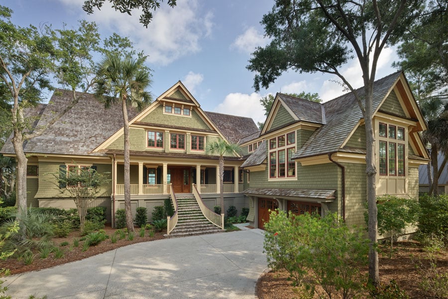 large, two-story Craftsman-style bungalow surrounded by lush greenery and tall trees. The house features a steeply pitched roof with shingles, multiple gables, and an inviting front porch with a wide staircase leading up to the entrance. The exterior is a mix of wood and stone, with numerous windows allowing natural light to pour into the home.