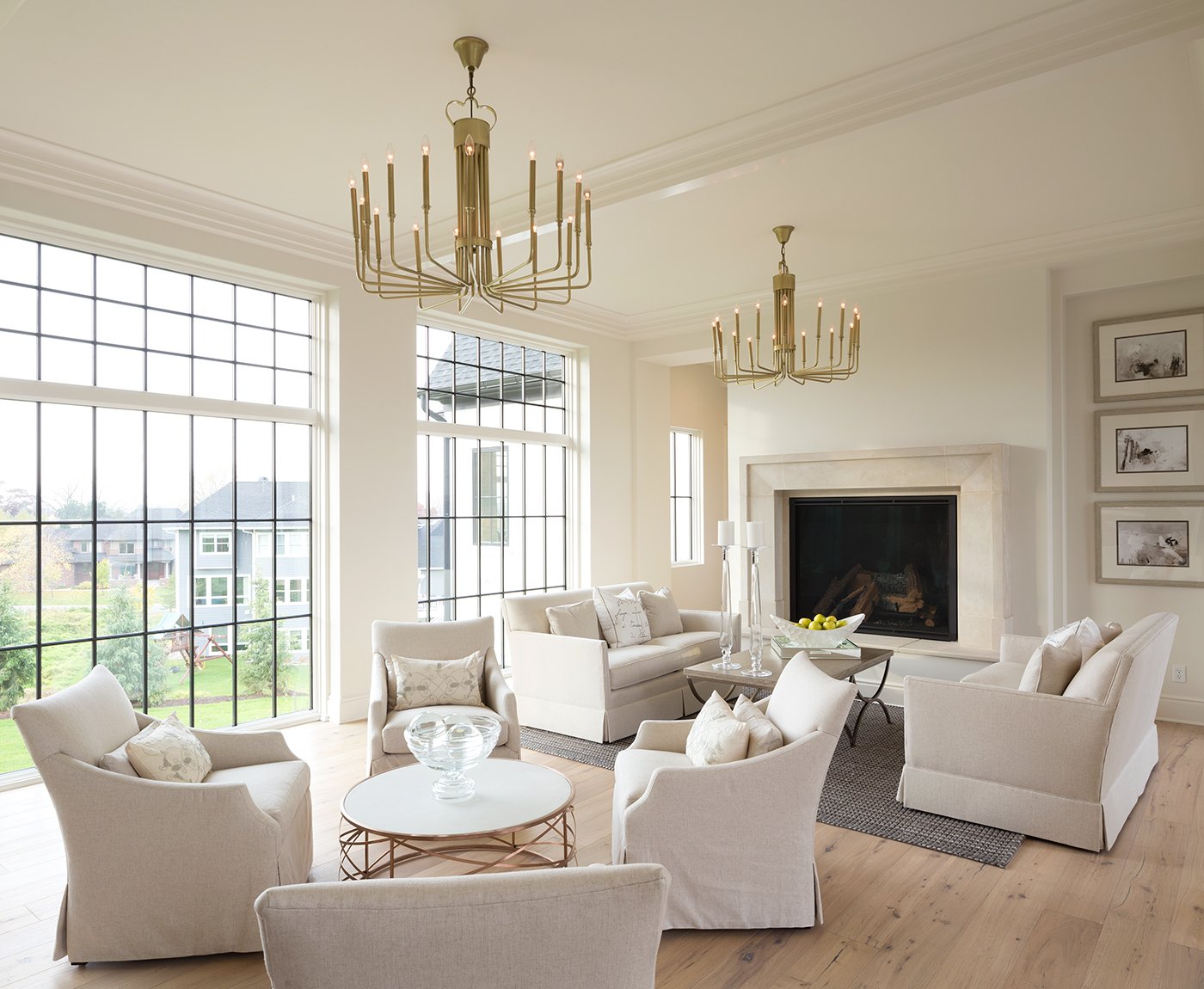 A large living room with white chairs and walls, gold chandeliers and a wall of casement and transom windows with rectangular black grilles. 
