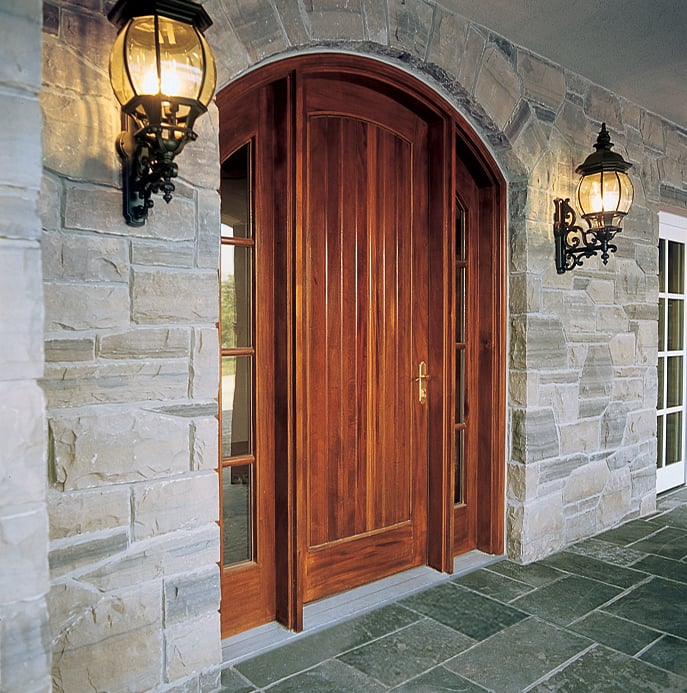 A dark plank door with brass hardware set inside a gray stone wall with sidelights and sconces.