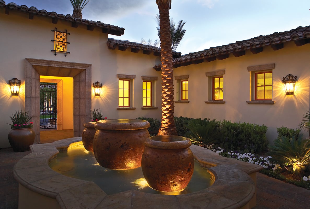 The exterior of a Spanish-style home at dusk with a fountain, palm trees, light stucco walls and casement windows with rectangular grilles.