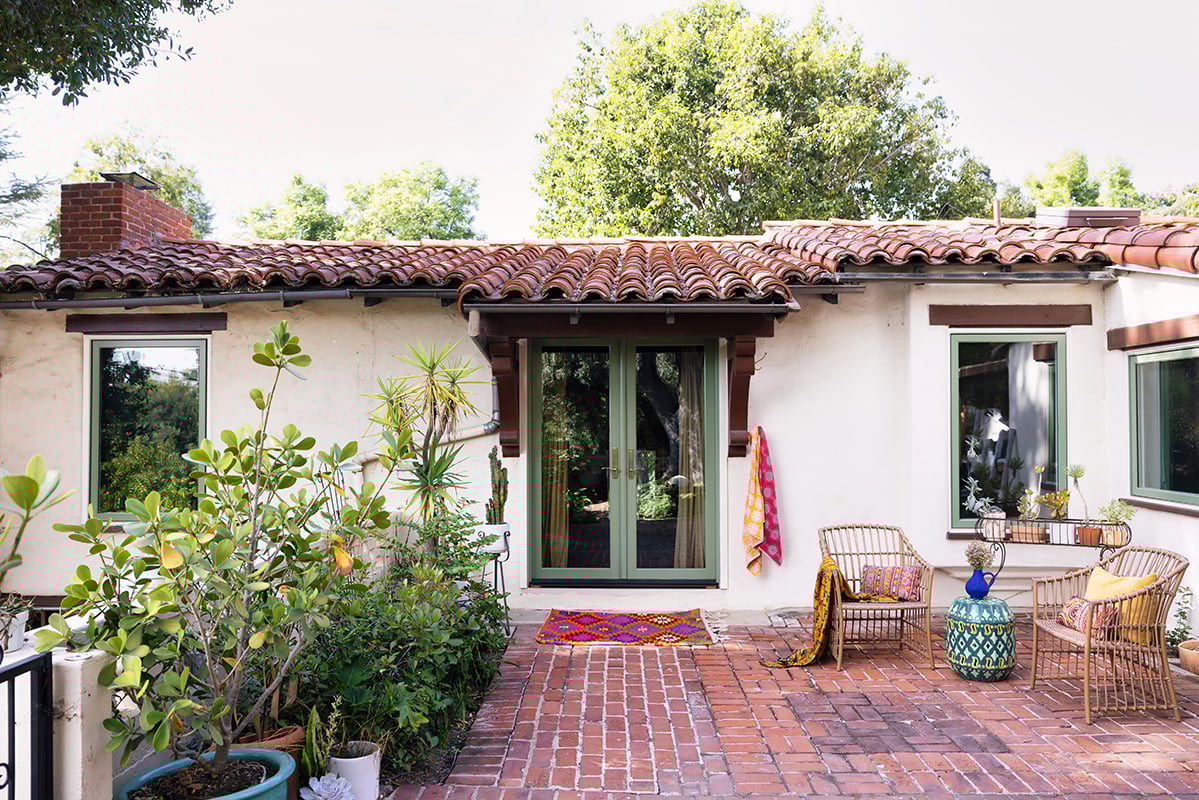 A brick patio living area outside a home with low pitched red tile roofs, white stucco, hinged patio doors and casement windows with green frames.