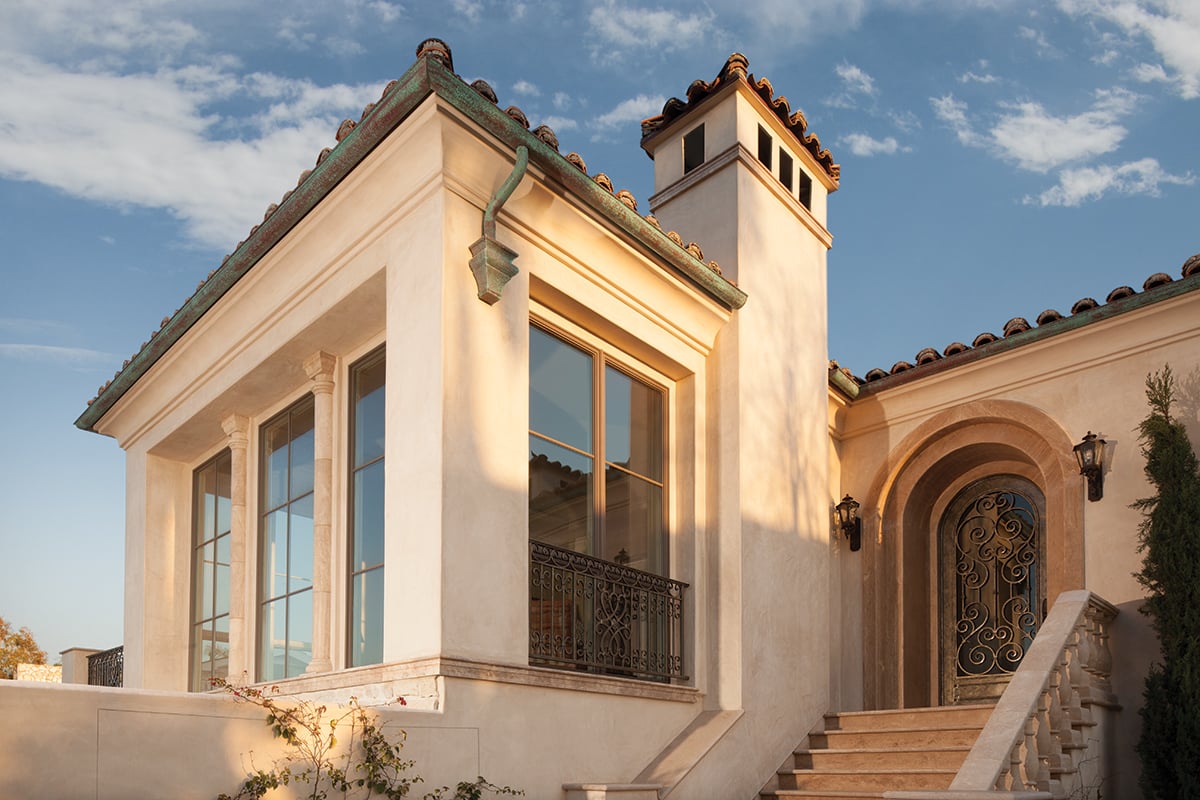 A white stucco Spanish-style home with floor-to-ceiling windows, tile roof, copper gutters, and wrought-iron details, including at the arched front door.