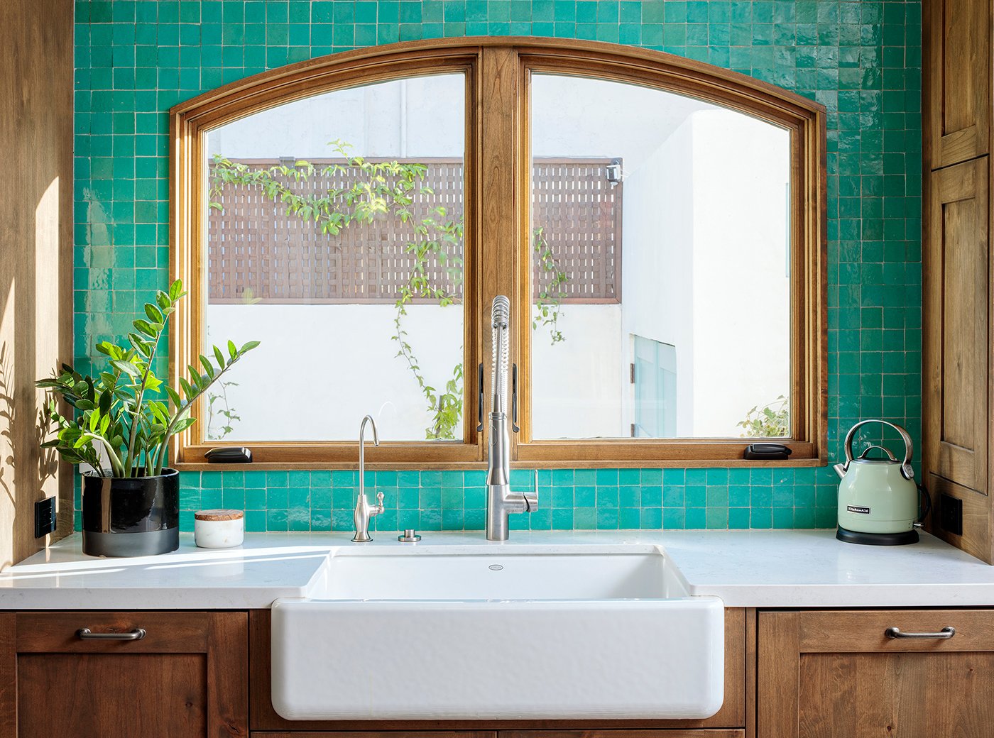 A white kitchen farm sink with turquoise tile backsplash and casement windows with an arch top and light wood frames