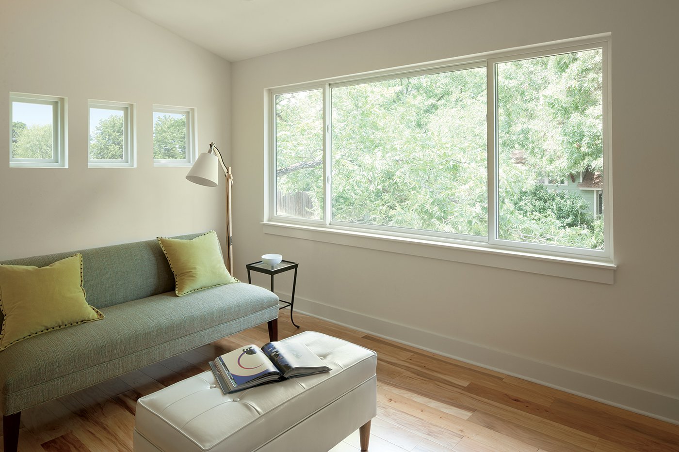 A living space with white walls and hardwood floors featuring a large 100 Series Gliding Window on one wall and three small picture windows on the other wall
