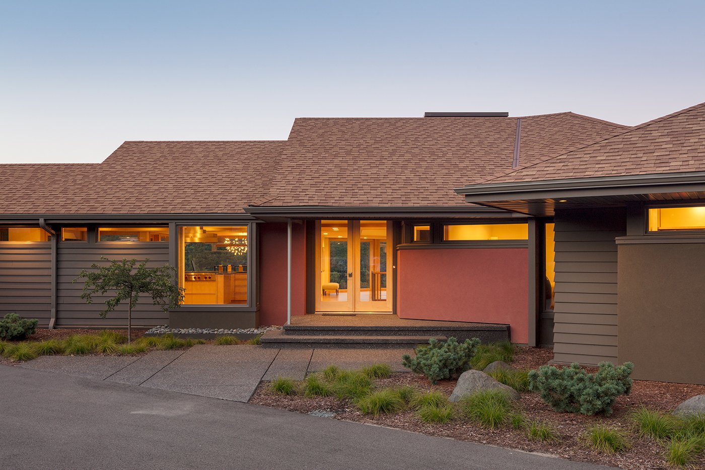 A Ranch home with earthy red exteriors and brown shingles lit up from the inside. 