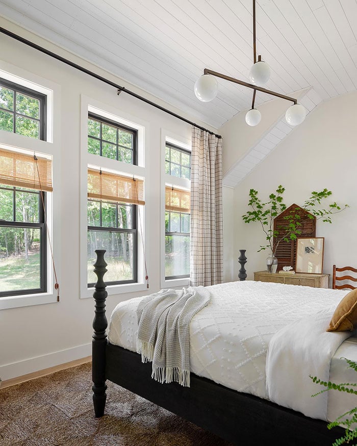 Two photos of a bedroom with warm white walls, black windows with modified colonial grilles, woven grass shades and a checked curtain, and a mix of heirloom wooden furniture.