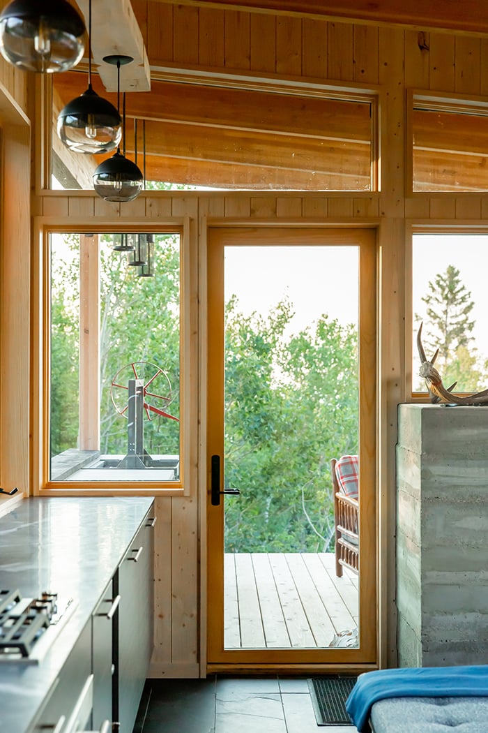 North Woods outdoor kitchen, including an indoor-out view through an all-glass hinged patio door and a second outdoor view of the kitchen featuring a grill, countertop, and cabinetry. 
