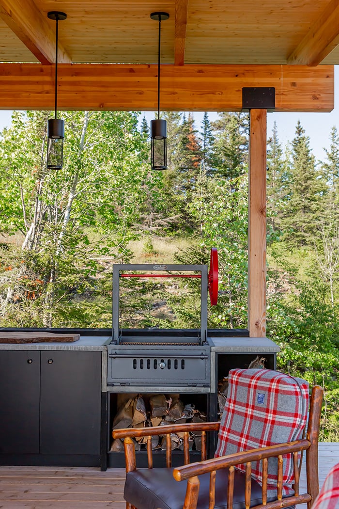 North Woods outdoor kitchen, including an indoor-out view through an all-glass hinged patio door and a second outdoor view of the kitchen featuring a grill, countertop, and cabinetry. 