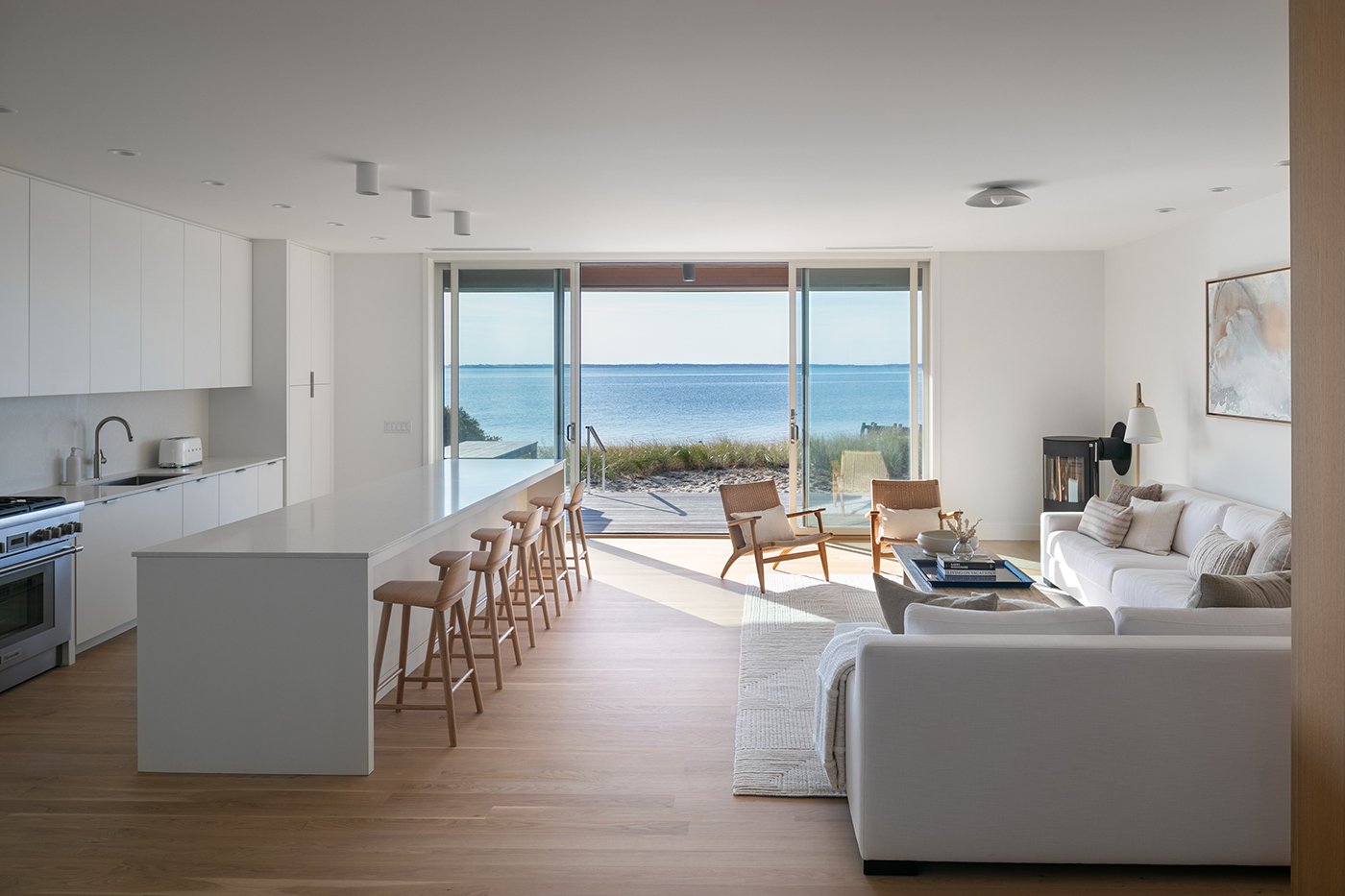 A shot of a living area of a home with white furniture and an open gliding patio door leading to the beach and a view of the water. 