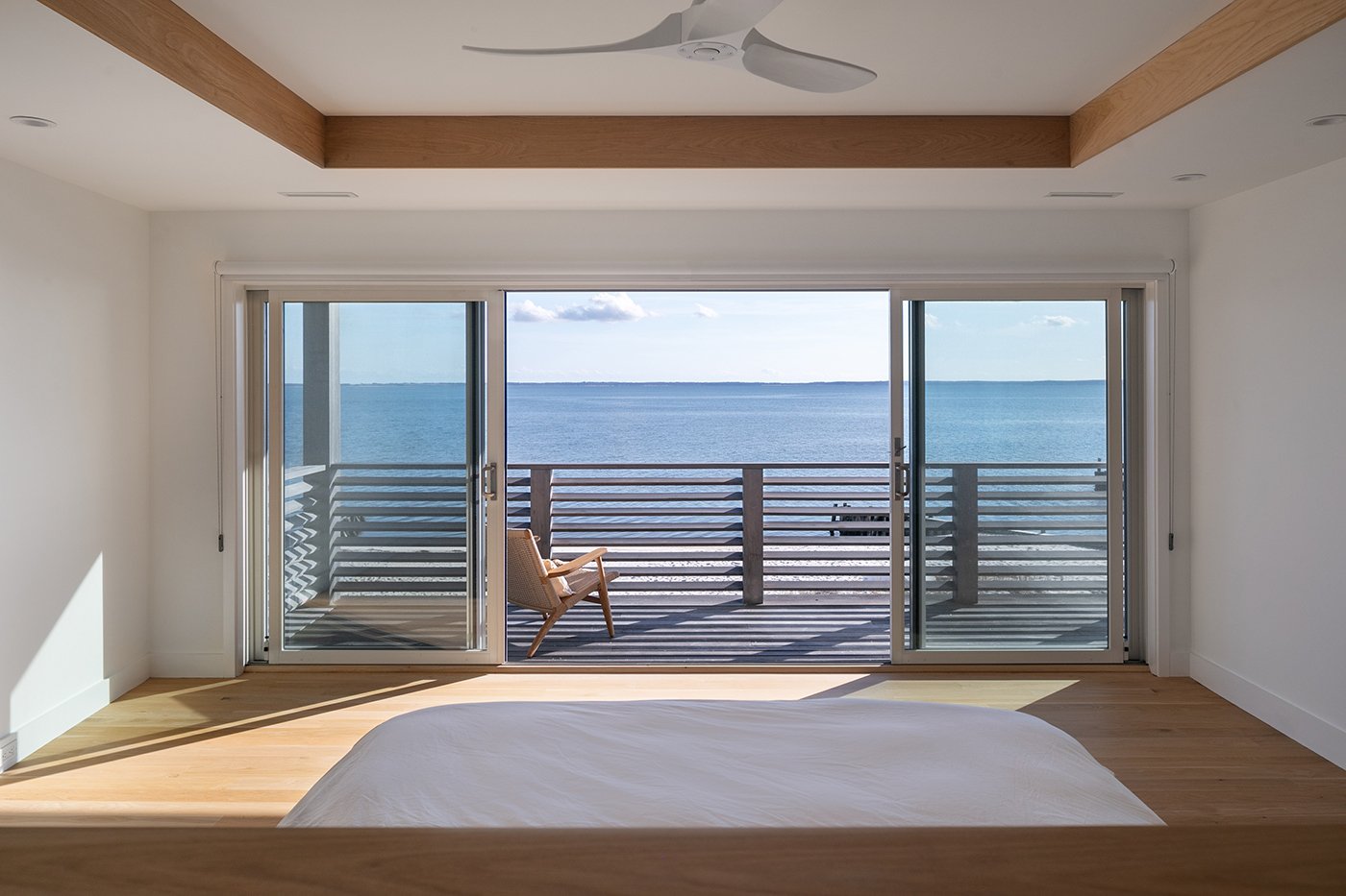 A bedroom with a wall of gliding patio doors and a wooden balcony with a view of the ocean. 