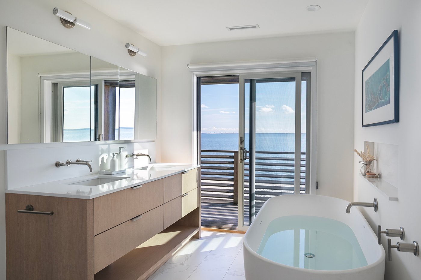 A bathroom with two sinks, a white tub, and a gliding patio door framing water views. 