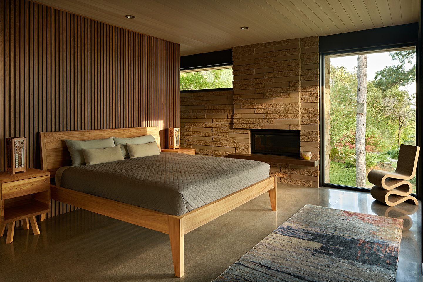 A bedroom with poured concrete floors, a stonework fireplace, and Andersen E-Series Picture Windows, including one that runs floor to ceiling.