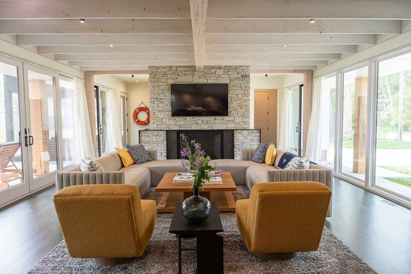 A living room with a stone fireplace, couches and chairs, and entire walls of windows and patio doors opposite each other.