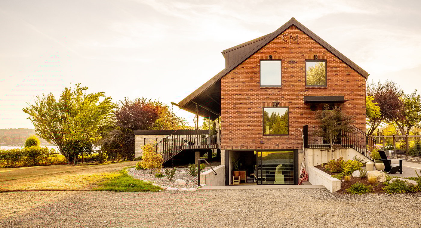 An exterior shot of a brick farmhouse with picture windows and roof overhang covering a waterfront deck.