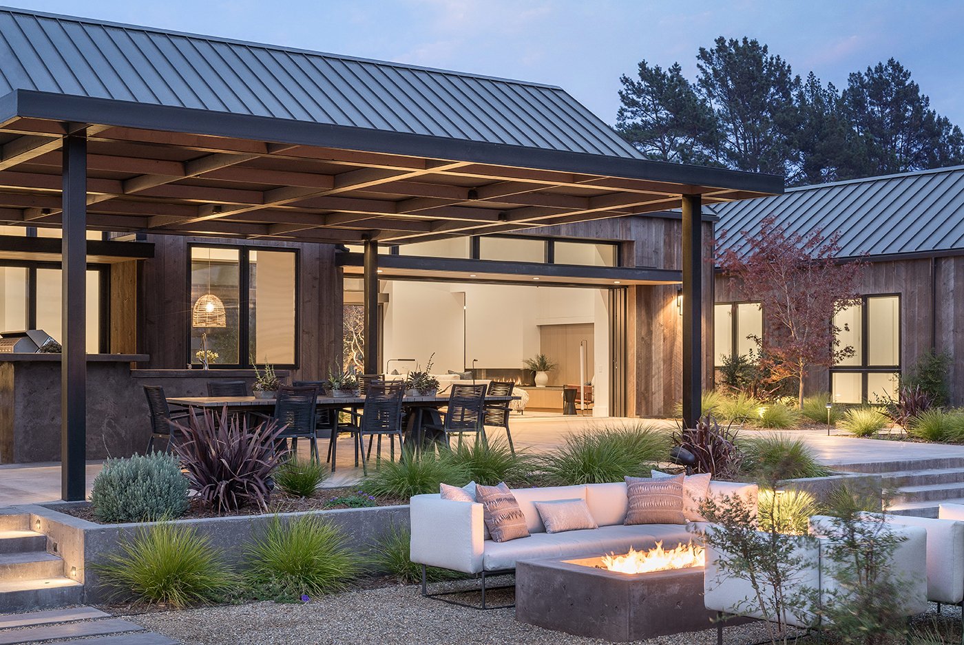 Andersen® E-Series windows and Liftslide Doors open into a backyard with an overhang, furniture and a crackling fireplace at dusk. 