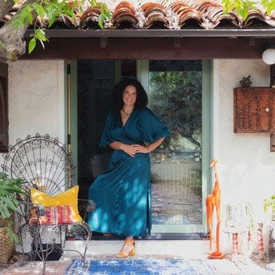 Justina Blakeney stands outside her stucco Spanish Colonial Revival home in front of an open E-Series Hinged Patio Door surrounded by potted plants, a chair, and a giraffe statue.