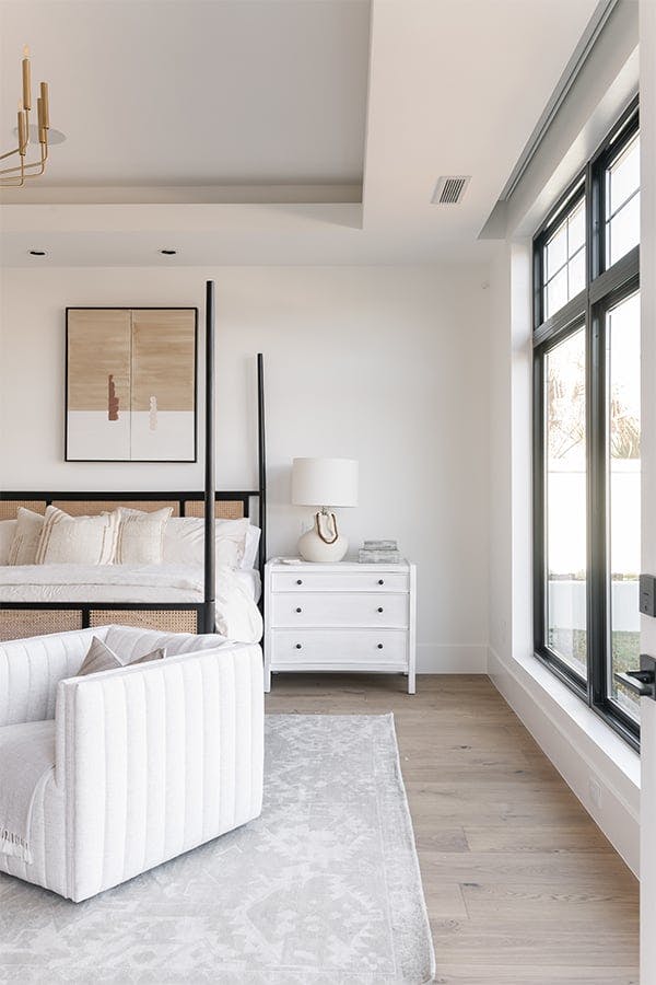 A white bedroom with a four-post bed, armchair, and nightstand has two tall black-framed windows with transoms above.
