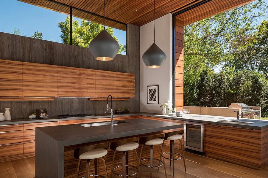 An open pass-through window connects an indoor kitchen with white oak cabinetry and marble countertops with an outdoor kitchen shaded by a large overhang. 