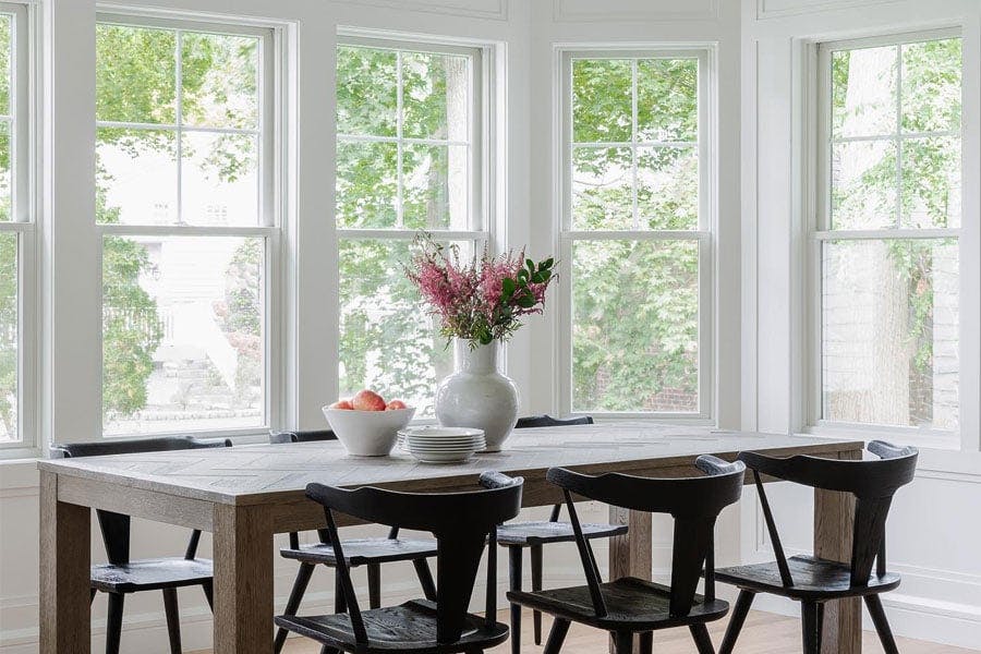  A breakfast room with a dining table and chairs is flooded with light from the three walls of windows with modified colonial grilles.