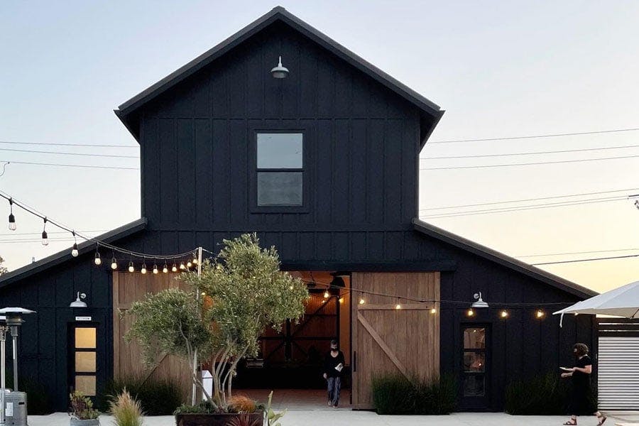 A black barn with a patio in front featuring a concrete firepit surrounded by chairs and potted plants and string lights overhead.