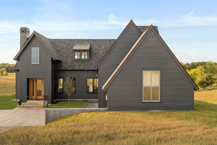 A charcoal farmhouse with a natural wood front door and blue-framed windows is set in a landscape of rolling prairie.